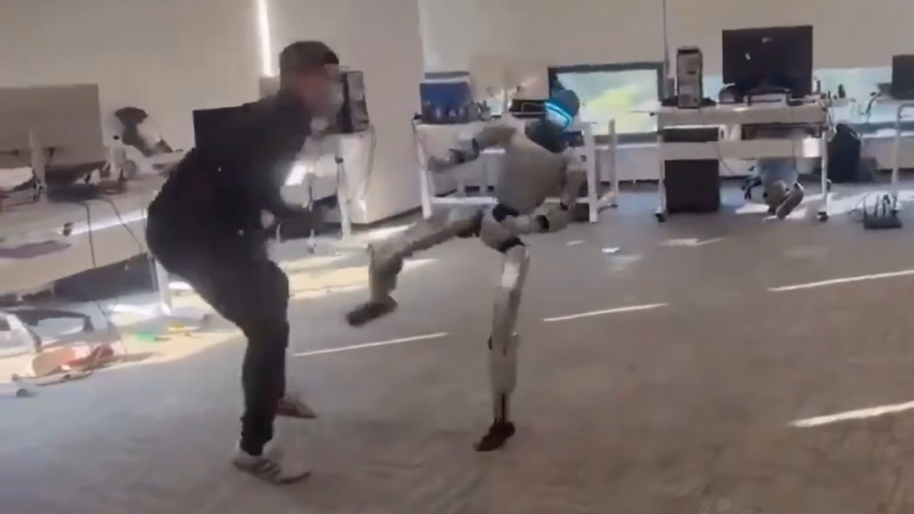 A silver humanoid robot performing a high kick towards a man in black clothing inside a bright office laboratory.